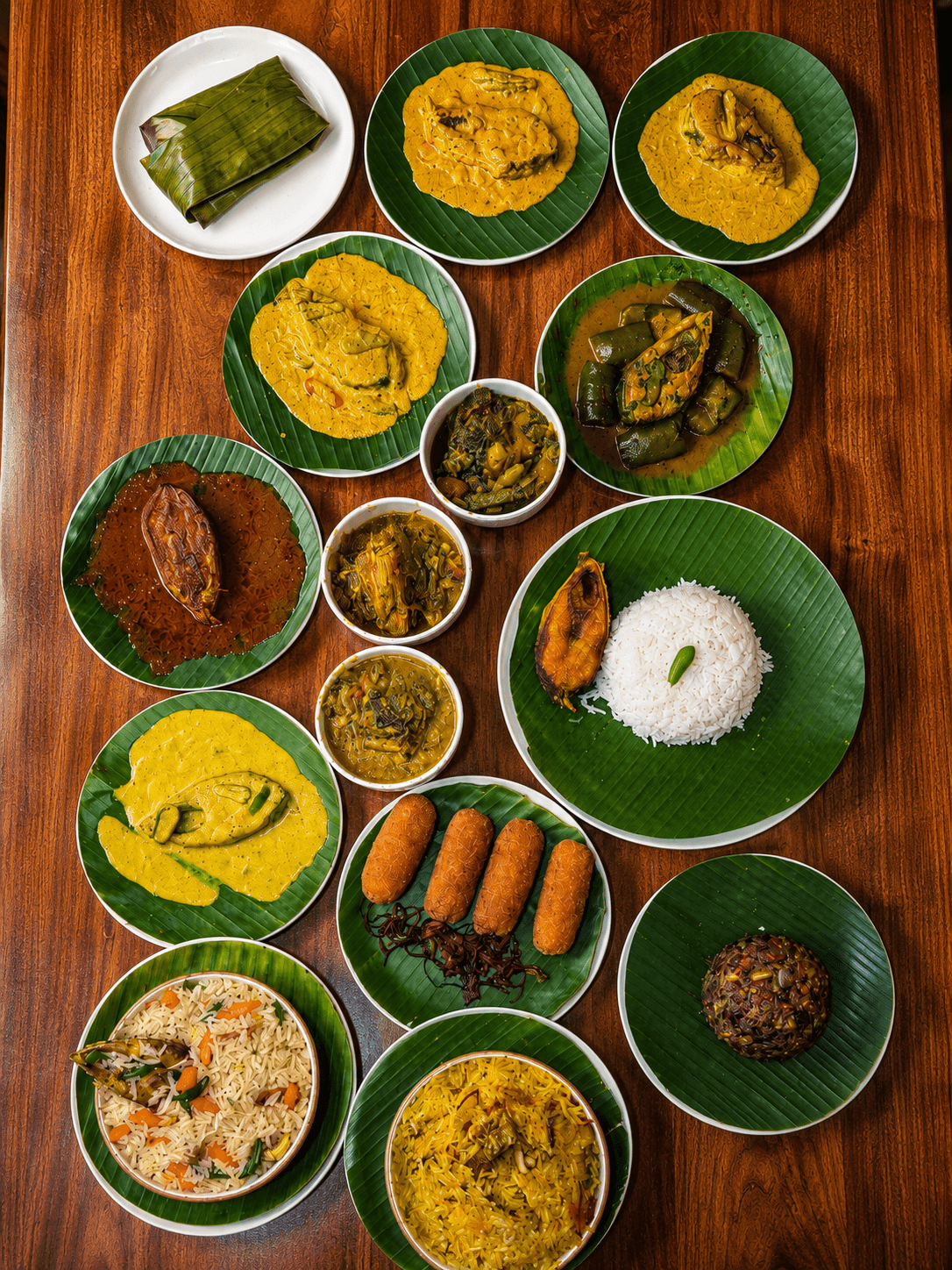 Traditional Bengali meal spread featuring various fish curries, rice, and snacks on green banana leaves.