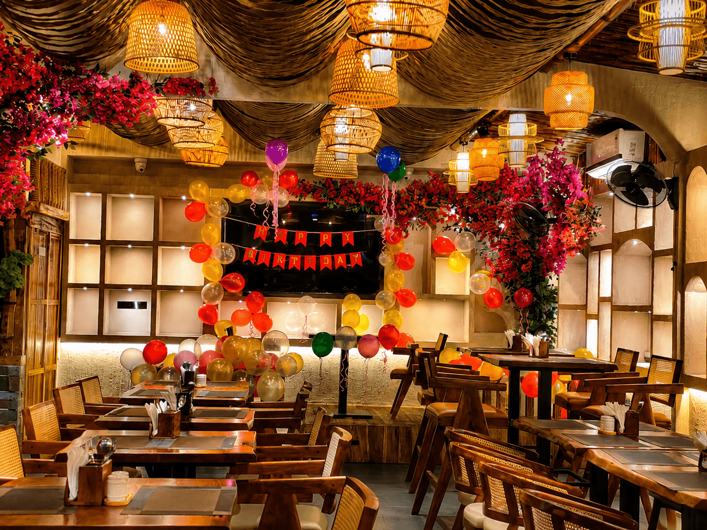 Festive restaurant interior decorated with colorful birthday balloons, a banner, floral accents, and woven lighting.
