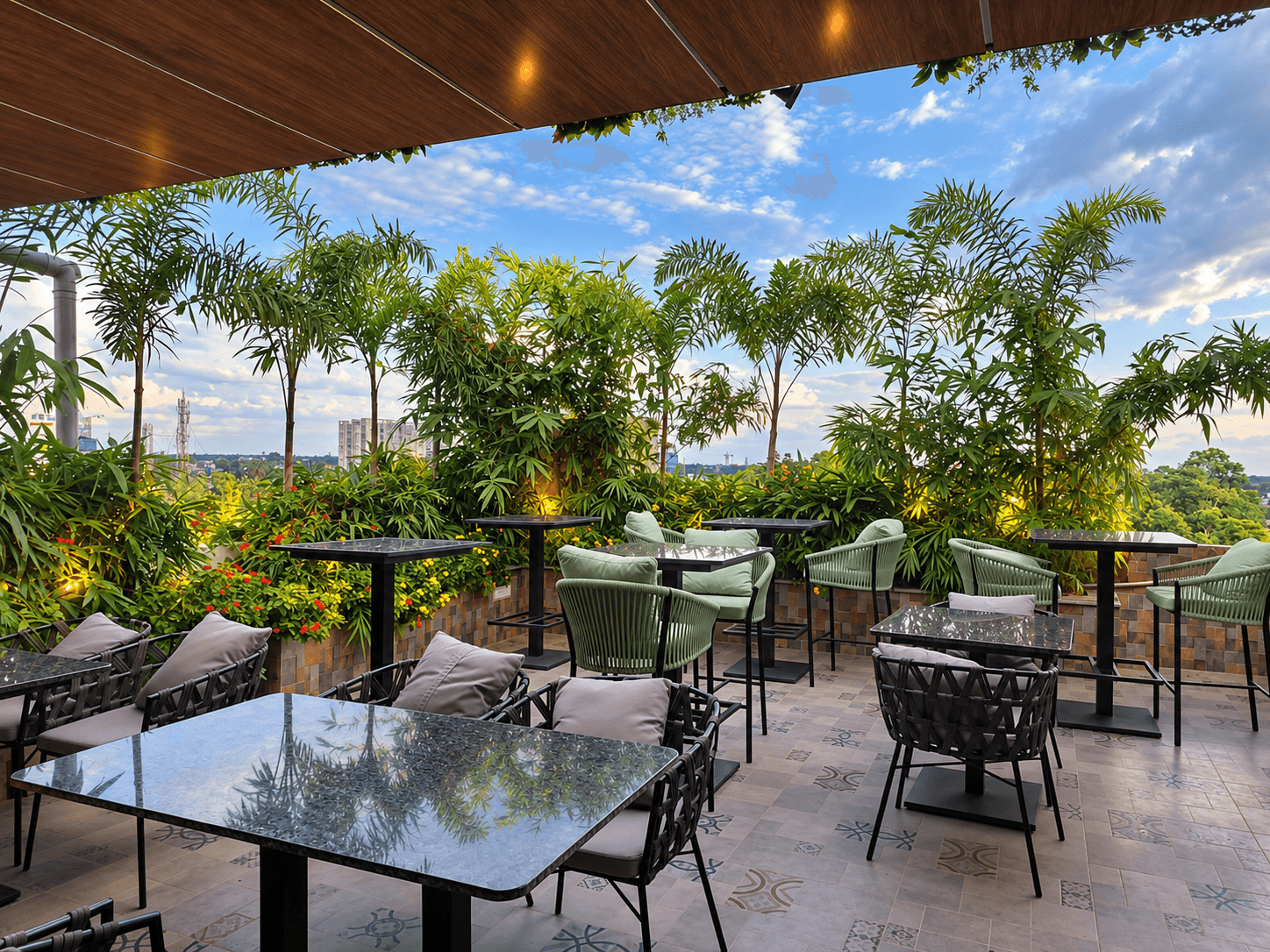 Rooftop patio with reflective tables, green woven chairs, and tropical plants under a cloudy sky.