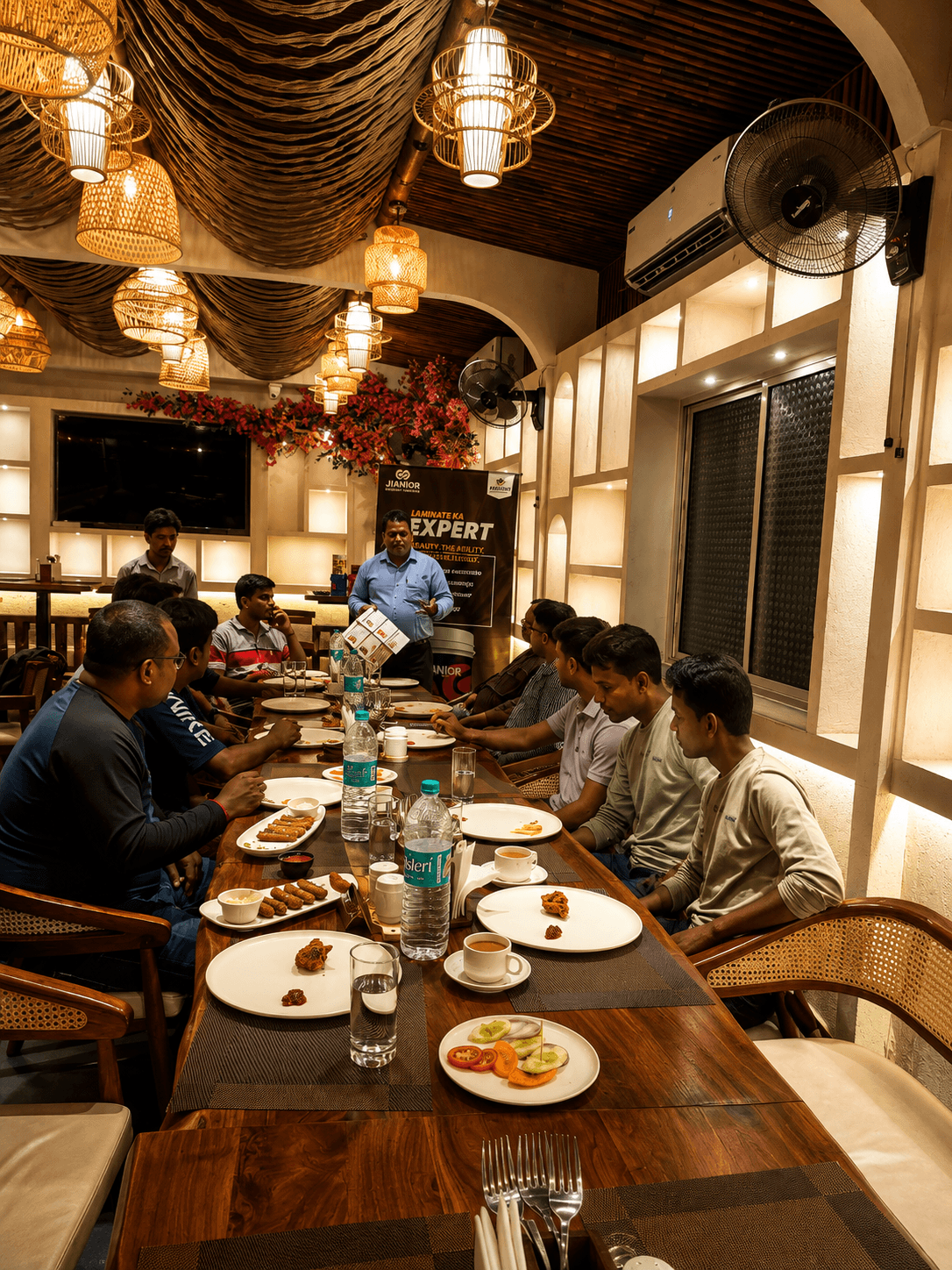 Men at a long restaurant table listening to a speaker under warm, decorative lighting.