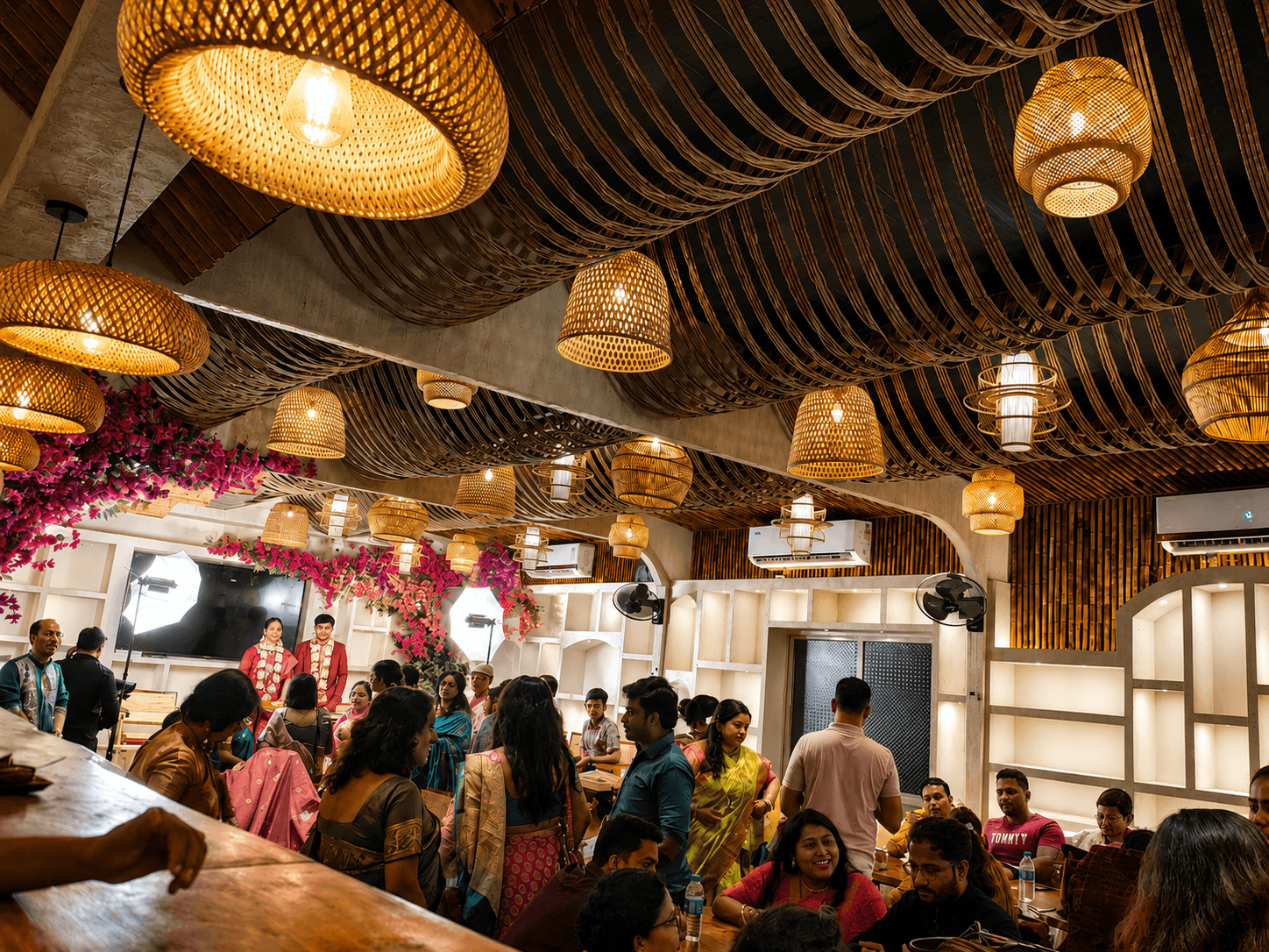Crowded restaurant interior featuring warm woven pendant lights and guests in colorful traditional South Asian attire.