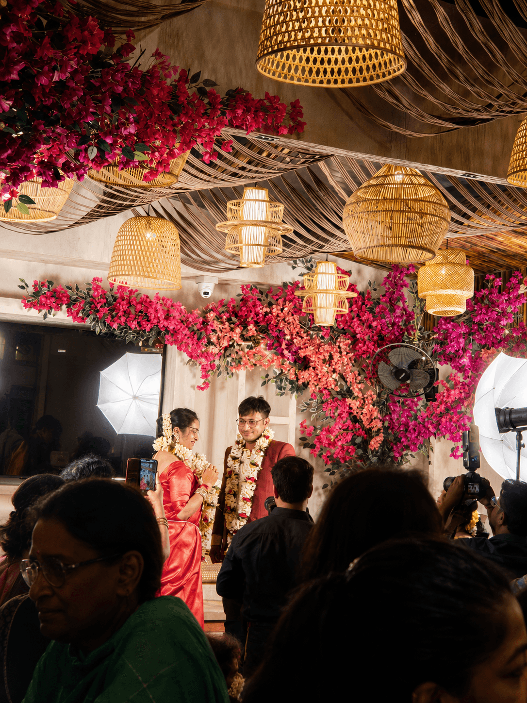A bride and groom smile under pink floral arrangements and woven wicker pendant lights.